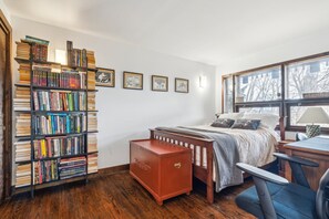Un dormitorio con una amplia estantería, una cama con cabecera de madera, un escritorio con silla y una ventana que da a árboles.