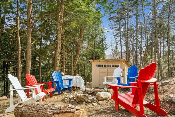 Un área boscosa con varias sillas Adirondack en colores rojo, blanco y azul, dispuestas alrededor de una pequeña estructura de madera.