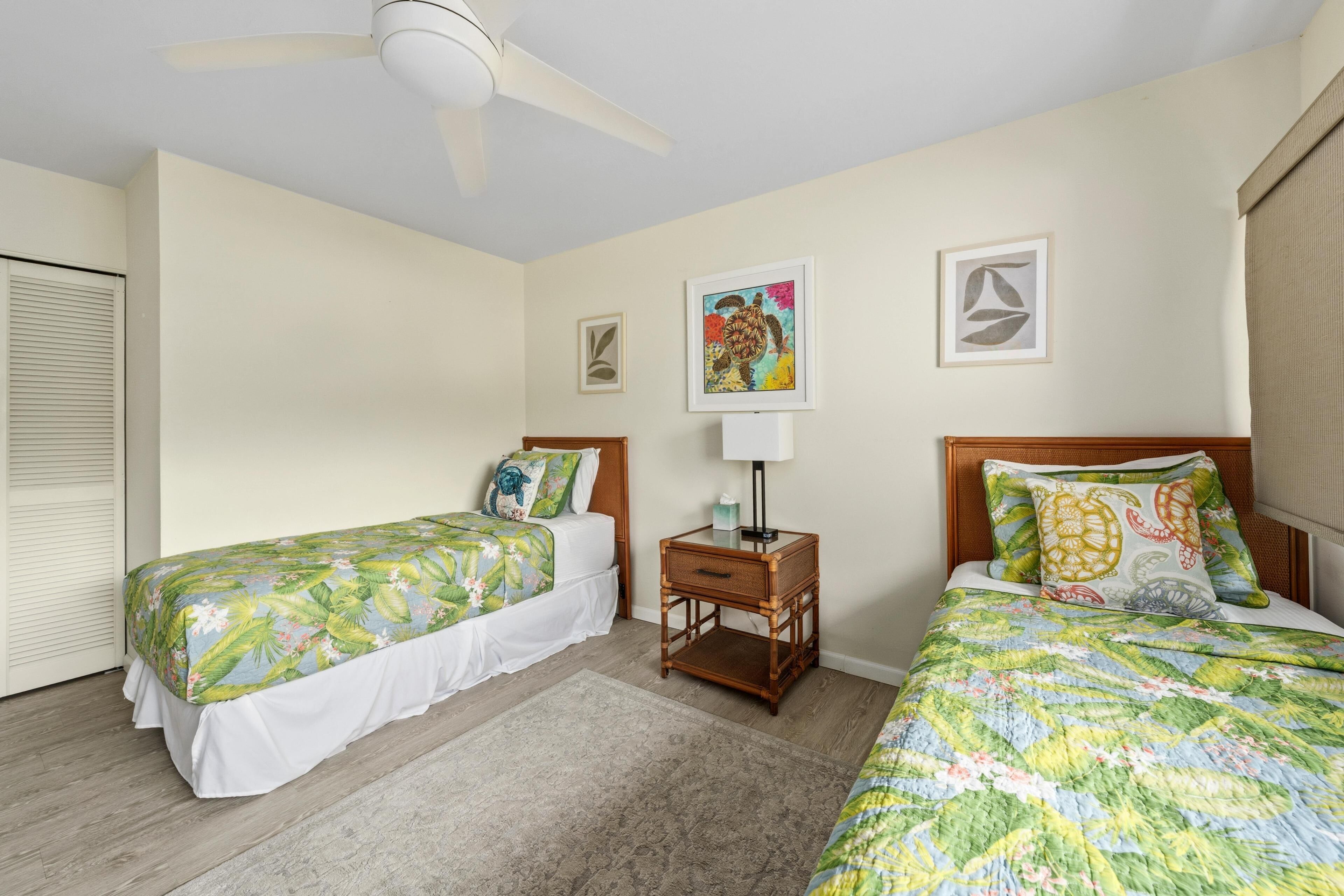 A hotel room with two single beds, a small table with a lamp, and a ceiling fan.