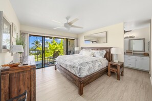 Un dormitorio con una cama grande, ventilador de techo, balcón con vista a palmeras y baño con espejo y armarios.