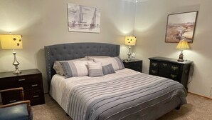 Ein Schlafzimmer mit einem geblatteten Kopfende, gestreifter Bettwäsche, zwei Nachttischen mit Lampen, einer Kommode und Kunstwerken an den Wänden.