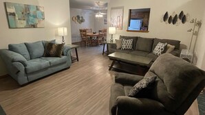 Ein Wohnzimmer mit Sofa, Sessel, Couchtisch, Essbereich mit Stühlen und einer durchgehenden Tür sichtbaren Küche.