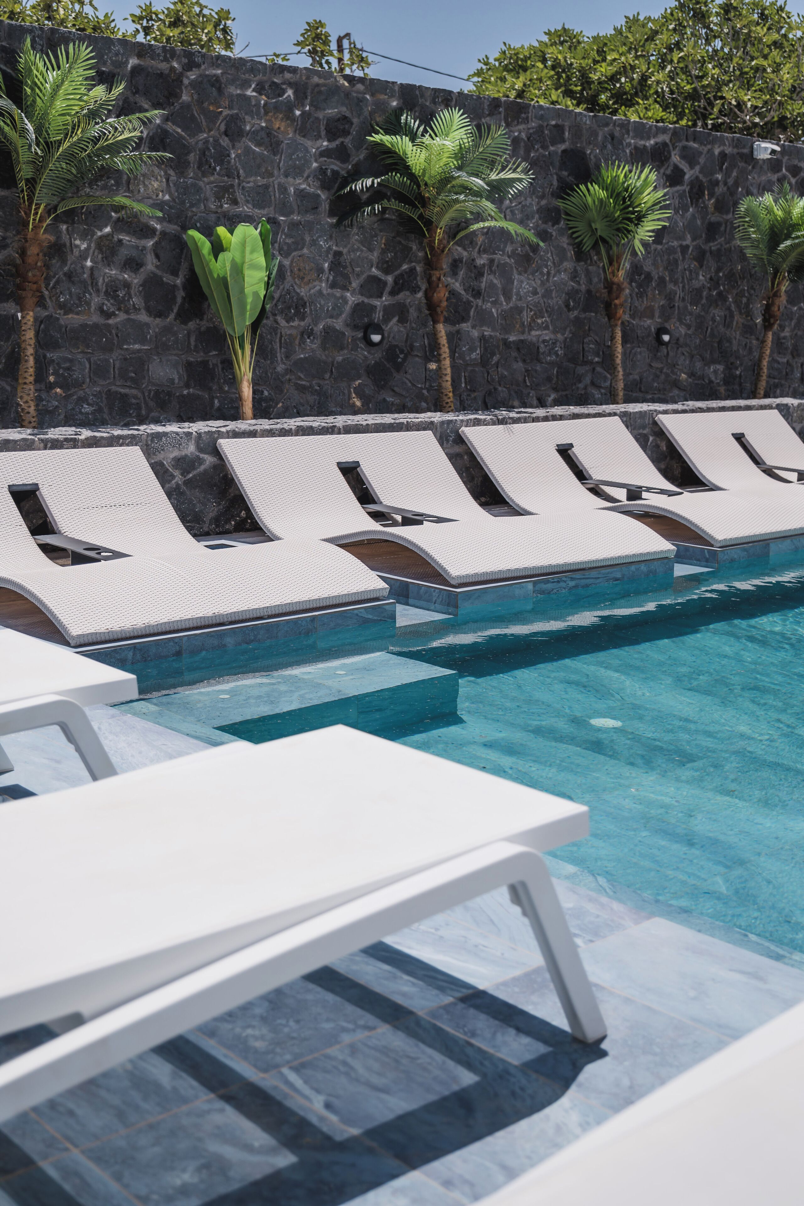 A poolside area with lounge chairs and palm trees against a stone wall.