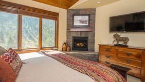 Un dormitorio con una cama grande, una chimenea de piedra, un televisor y vistas a un bosque a través de amplios ventanales.