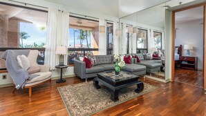 A spacious living room with a large sofa set, a coffee table, and a chair. There is a large window with curtains, a mirror, and a view of palm trees. - Includes Beach Gear!  332 Shores at Waikoloa (Waikoloa Beach Resort)