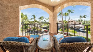 A balcony with wicker furniture and a glass table, overlooking a residential area with palm trees and houses. - Includes Beach Gear!  332 Shores at Waikoloa (Waikoloa Beach Resort)