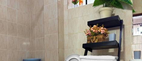 A modern bathroom with a toilet, a blue basin, a shelf with towels, a potted plant, and a window with flowers.
