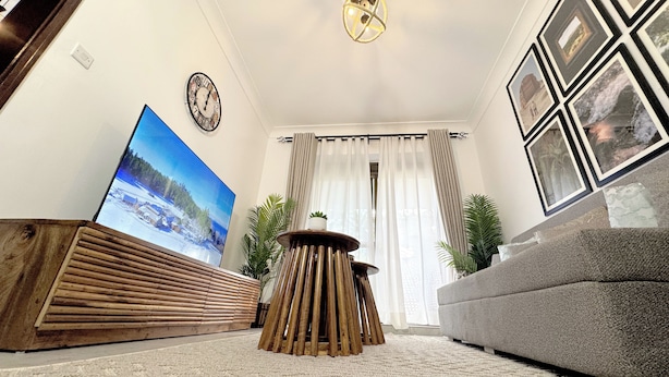 A living room with a large flat-screen TV, a grey sofa, a wooden coffee table, and a patterned rug. - The Home Kololo (Kampala)