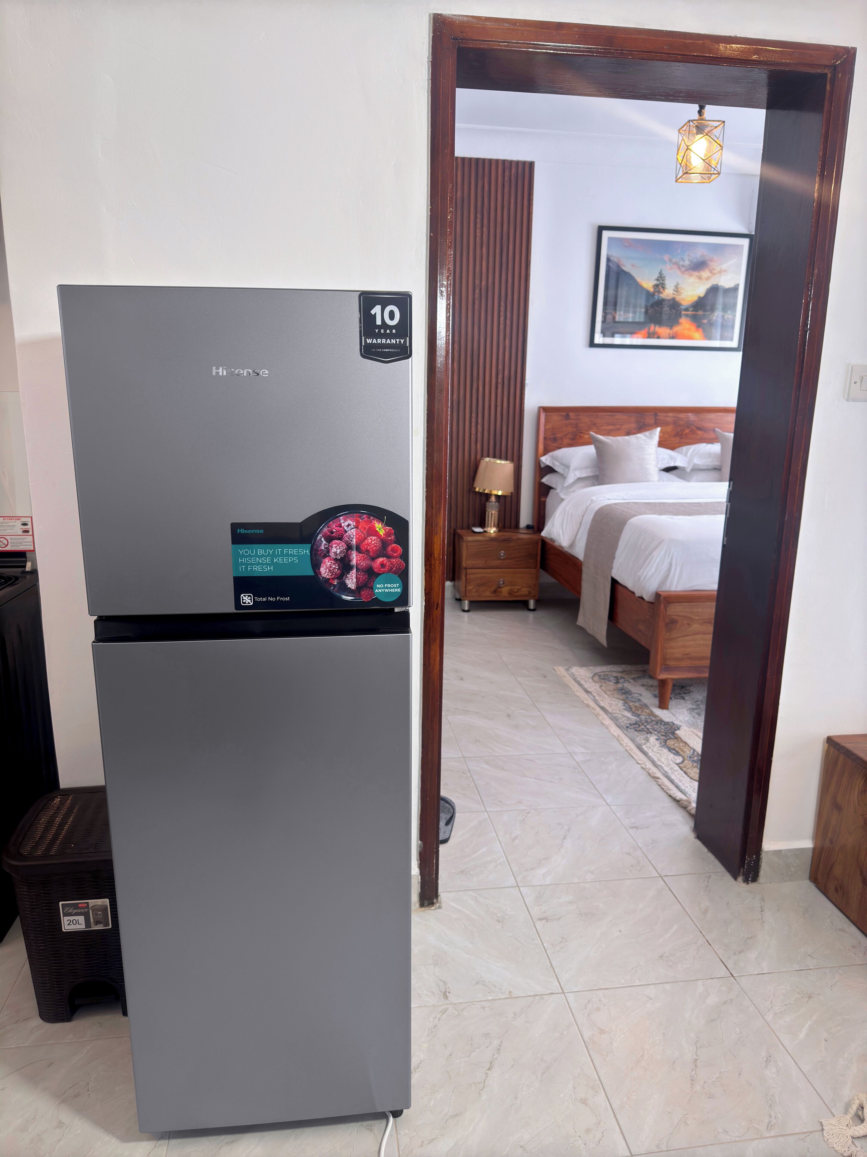 A compact, modern hotel room with a grey Hisense refrigerator, a wooden bed, a nightstand with a lamp, and a framed picture on the wall.