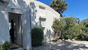 Un bâtiment blanc avec une porte ouverte donnant sur un couloir, entouré de gravier et de plantes.