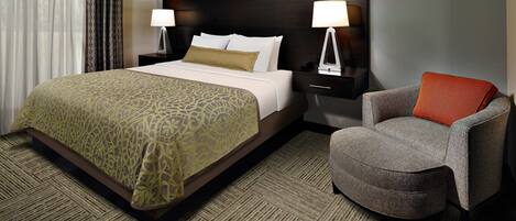 A hotel room with a large bed, two bedside tables with lamps, a grey armchair, and a patterned carpet.