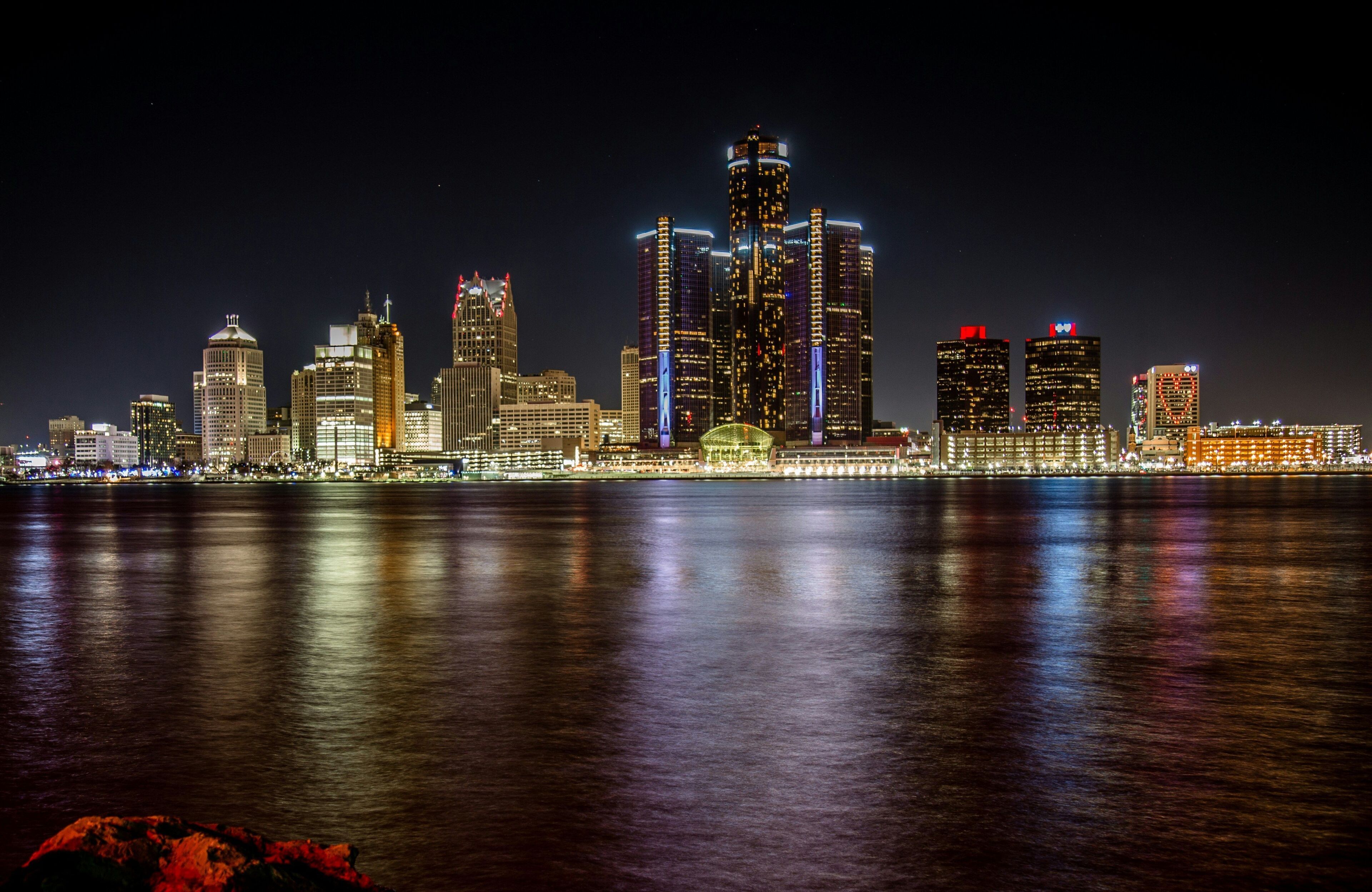 A nighttime city skyline with illuminated buildings reflecting on the water.