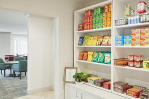 A kitchen with shelves stocked with various food items, a dining area with chairs, and a framed picture on the counter.