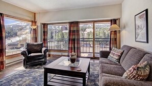 A living room with a sofa, armchair, and coffee table, large windows with curtains, and a view of a mountainous landscape. - Huge Slope-Side Condo (Breckenridge)