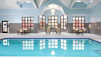 Indoor swimming pool with a tiled pool deck, large windows, and lounge seating.
