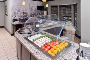 Zona de bufé con gran variedad de alimentos, entre ellos frutas y productos lácteos, en un ambiente moderno con vitrina de vidrio.