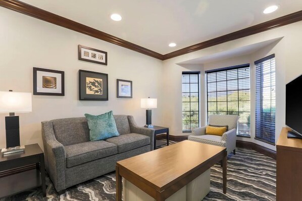 A hotel room with a gray sofa, a wooden coffee table, a TV, and framed artwork on the wall.