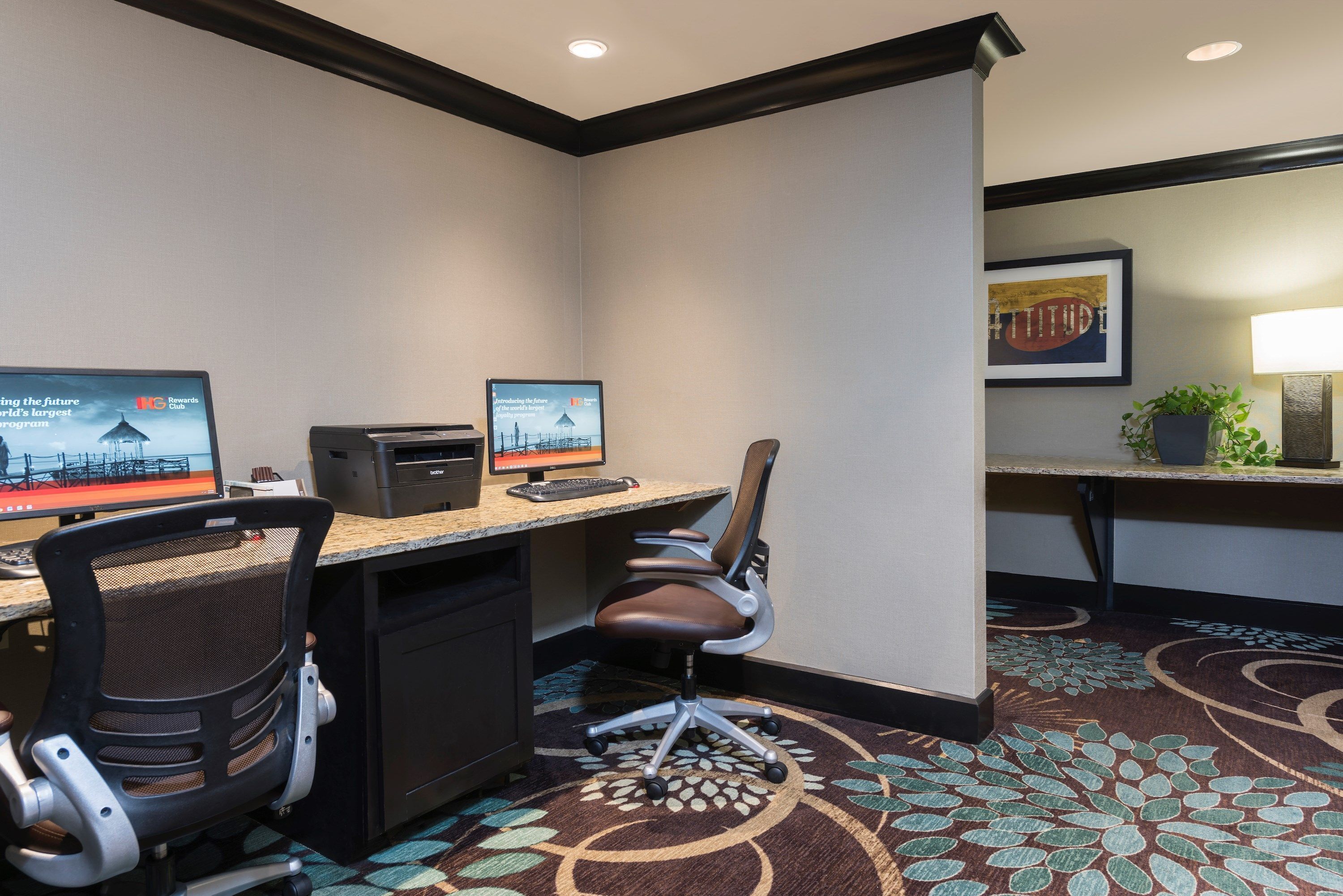 A hotel room with a desk, two monitors, a chair, a small desk lamp, and a decorative rug.