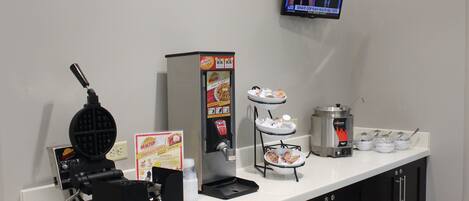 Een self-service ontbijtstation met een pannenkoekenijzer, koffieapparaat en een televisie die nieuws laat zien.