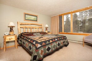 A bedroom with a wooden headboard, a quilt, a bedside table with a lamp, a chair, and a window with a view of trees. - Condo Along Snake River | Hot Tub Access | Shuttle (Keystone)