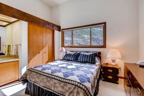 Ein Schlafzimmer mit Bett, Nachttisch, Kommode, Fenster mit Blick auf Bäume und einem Holzbalken an der Decke.