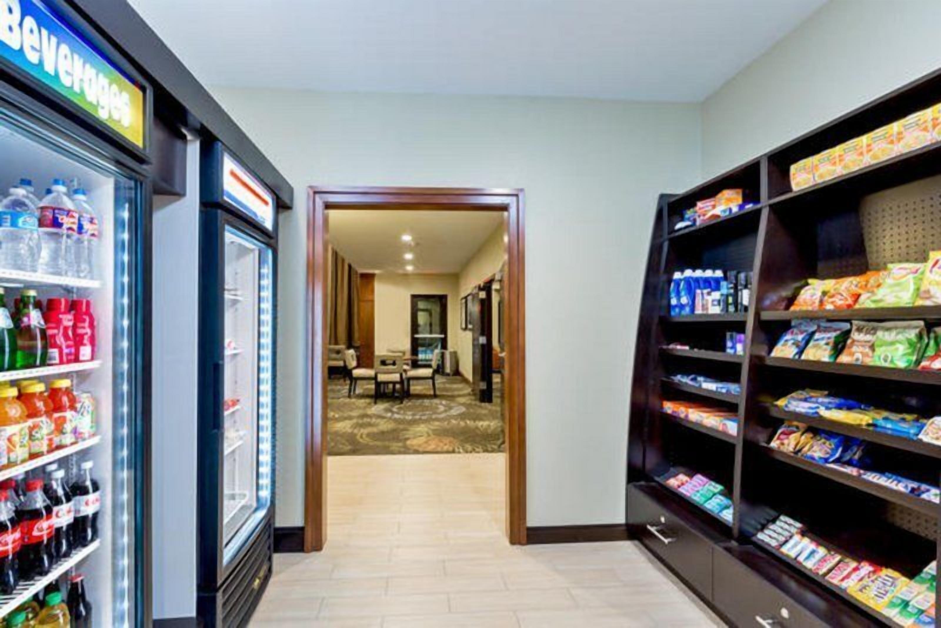 A hotel lobby with a beverage refrigerator, snack shelves, and a carpeted area leading to a room.