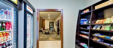 A hotel lobby with a beverage refrigerator, snack shelves, and a carpeted area leading to a room.