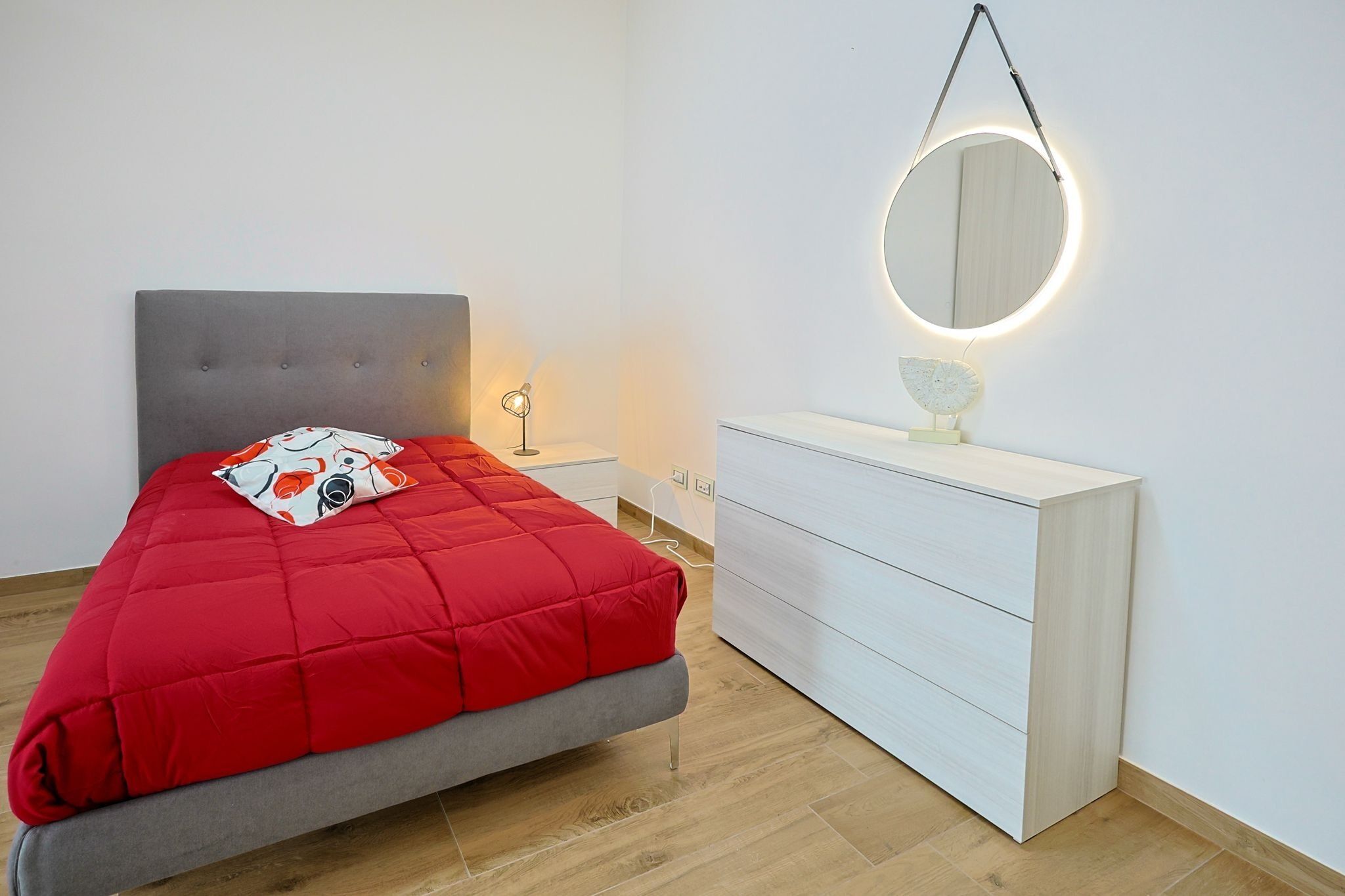 Ein Schlafzimmer mit einem grau gepolsterten Bett, einer weißen Kommode, einem runden Spiegel mit Beleuchtung und einem kleinen Tisch mit Lampe.