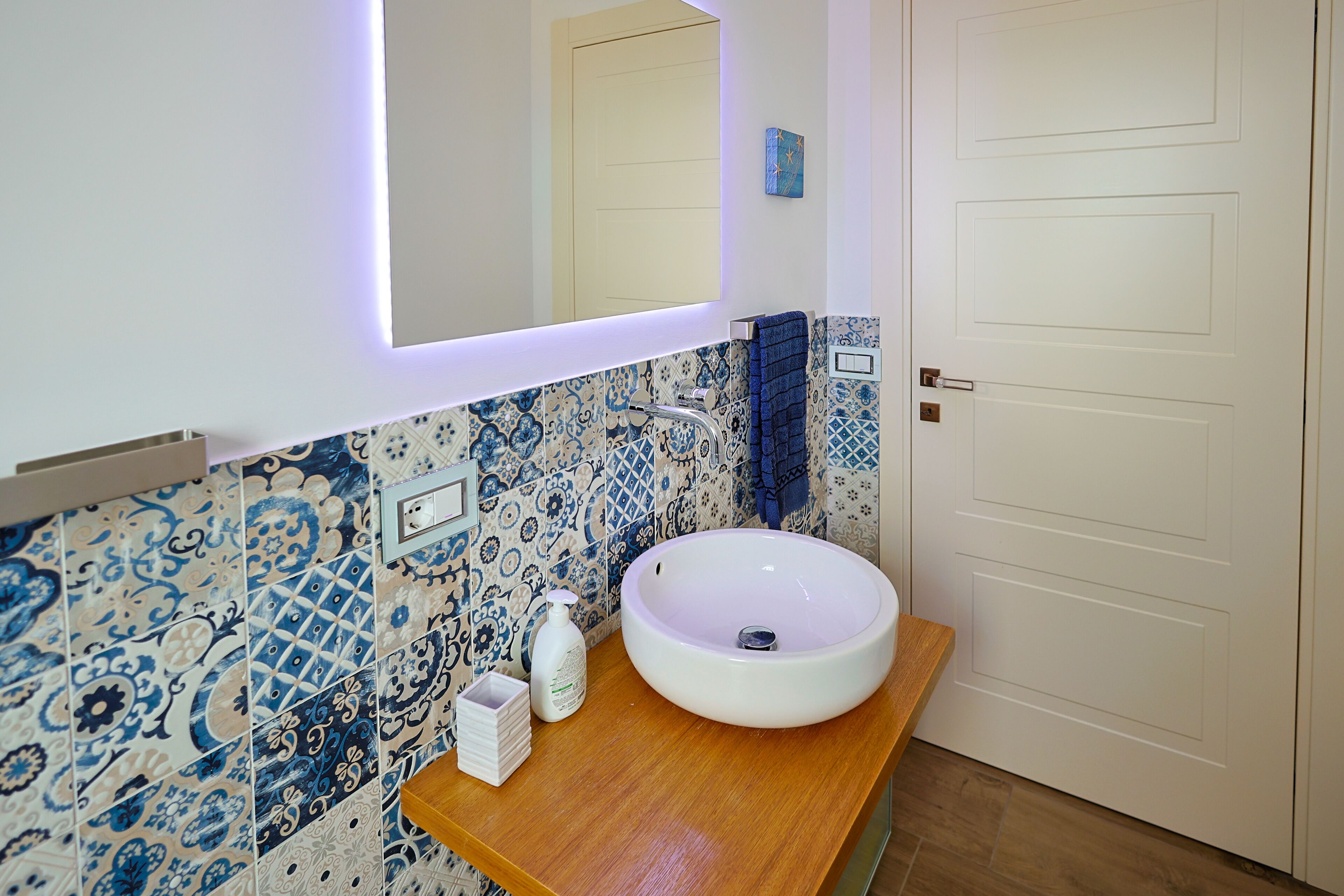 A bathroom with a white sink, blue patterned tiles, a wooden countertop, a mirror with a light, and a door.