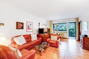 A living room with a red sofa, a glass coffee table, and a television. There is a window with a view of trees and a balcony.