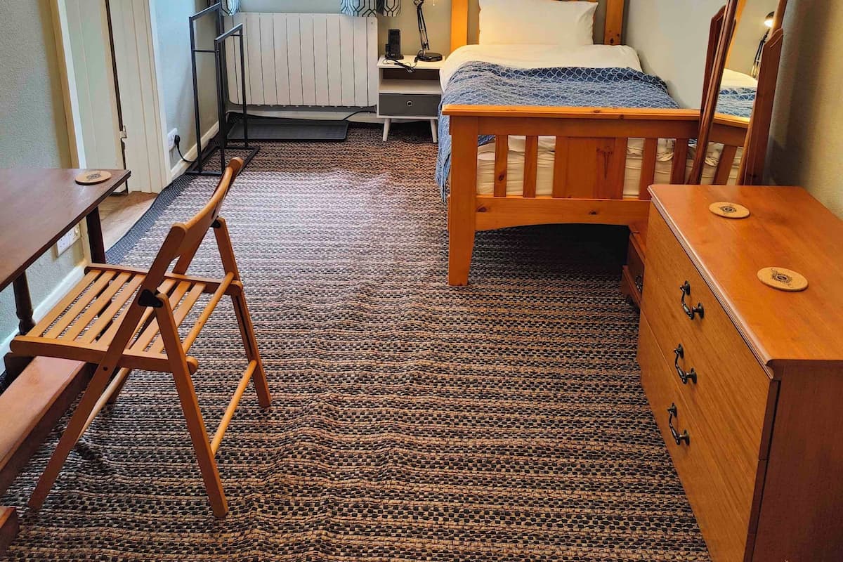Ein Schlafzimmer mit einem hölzernen Bett, einem Nachttisch, einer Kommode, einem Stuhl, einem Fenster mit Vorhängen und einem Heizkörper.