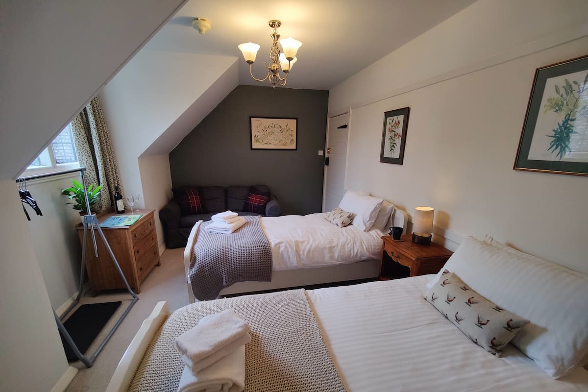 Ein Schlafzimmer mit zwei Betten, einer Stehlampe, einem Nachttisch, einem Fenster mit Vorhängen, einer Pflanze und einer Deckenleuchte.