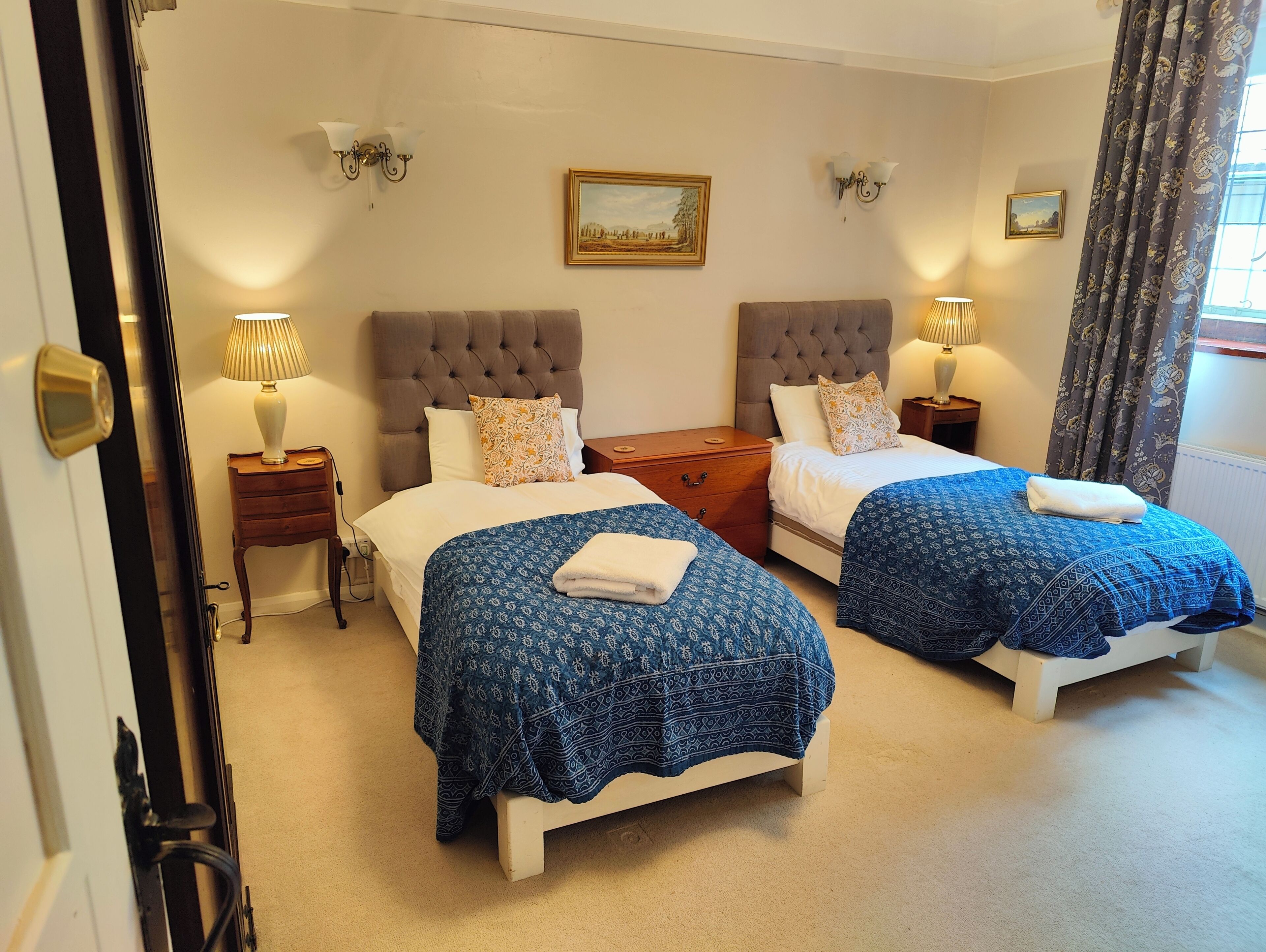 A hotel room with two beds, a painting on the wall, and a window with curtains.