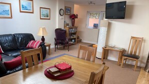A living room with a wooden dining table, chairs, a leather sofa, a bookshelf, a TV mounted on the wall, and a window with a view. - Seaside Retreat in Exmouth With Stunning Views (Exmouth)