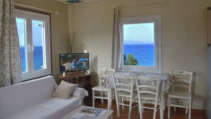 A living room with a sofa, a TV, a dining table with chairs, and a view of the sea through a window. - Aegean Bliss Residences Karfas (Chios)