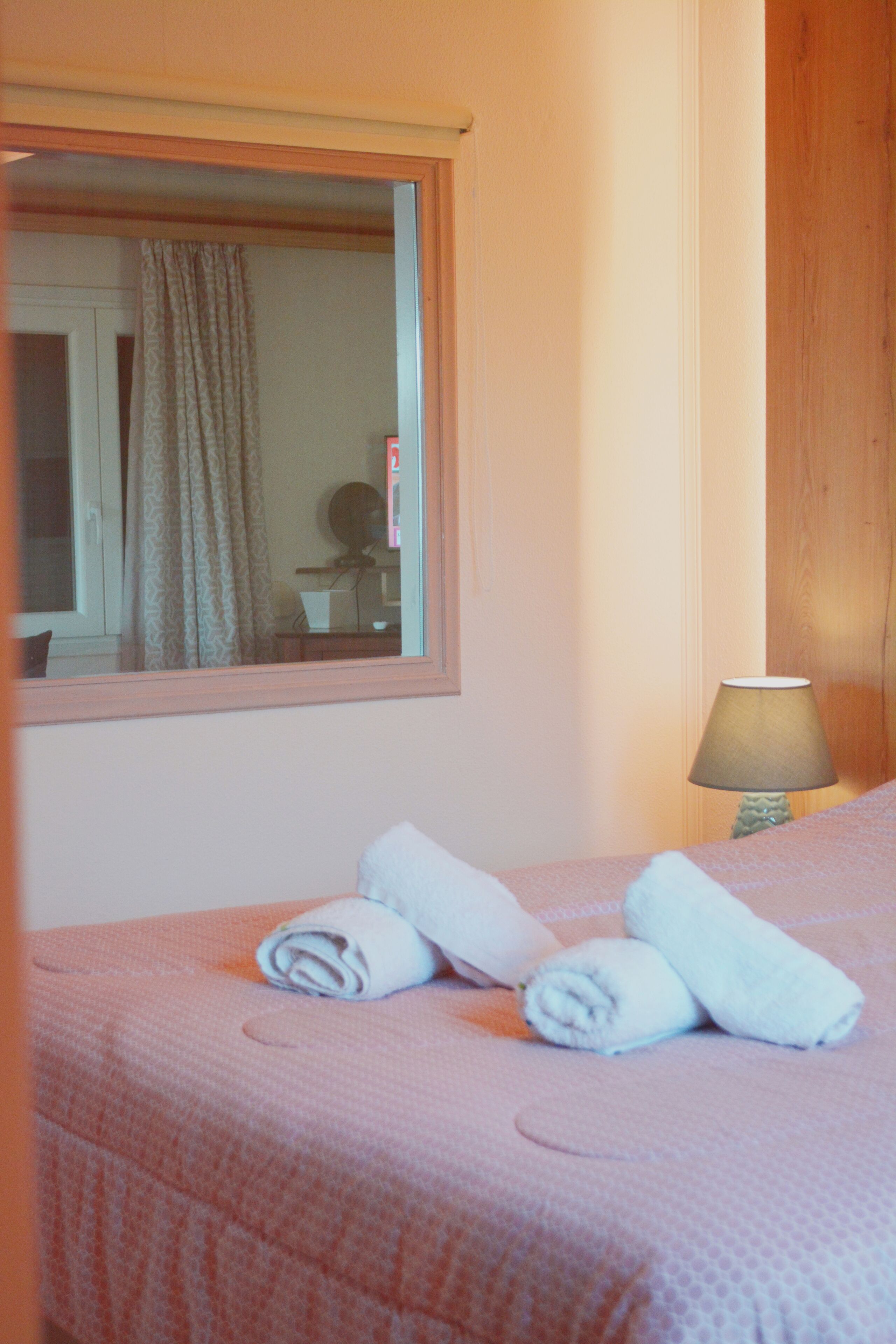 A neatly made bed with rolled towels, a window with curtains, and a lamp on a bedside table.