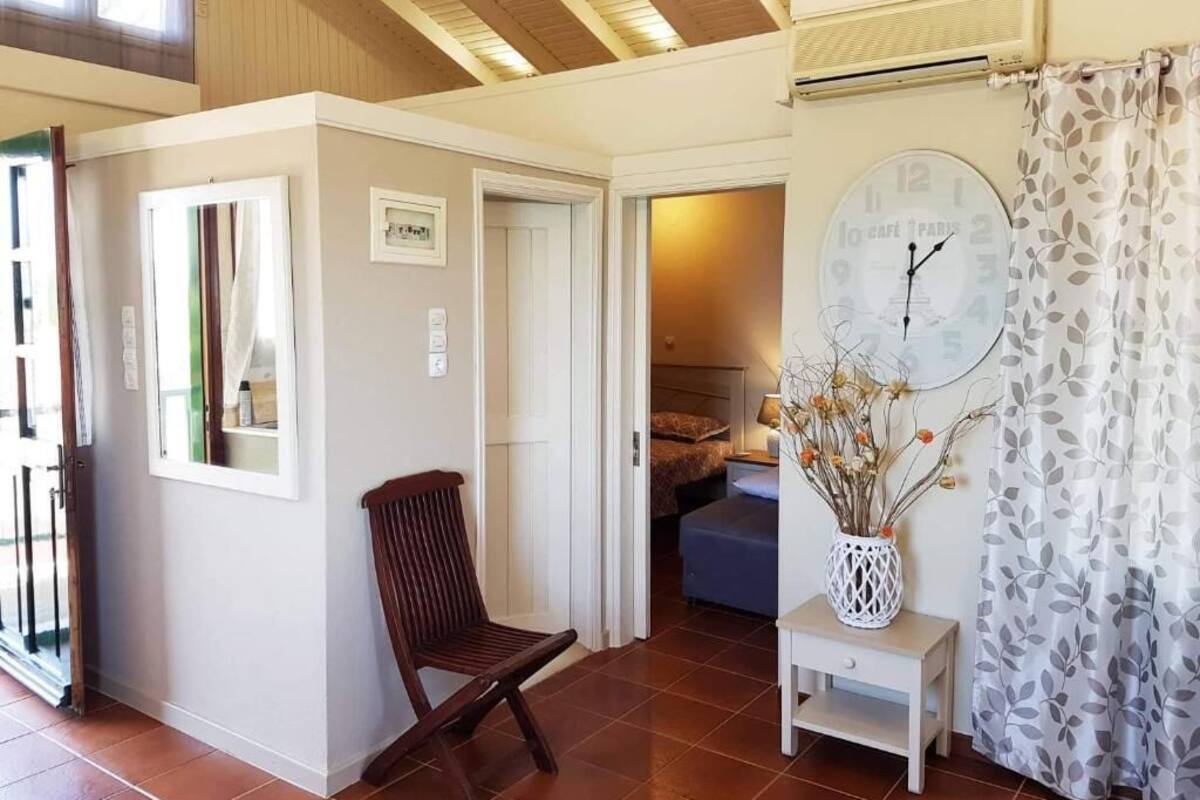 A room with a wooden ceiling, a clock on the wall, a chair, a small table with a vase, and a bed in the background.