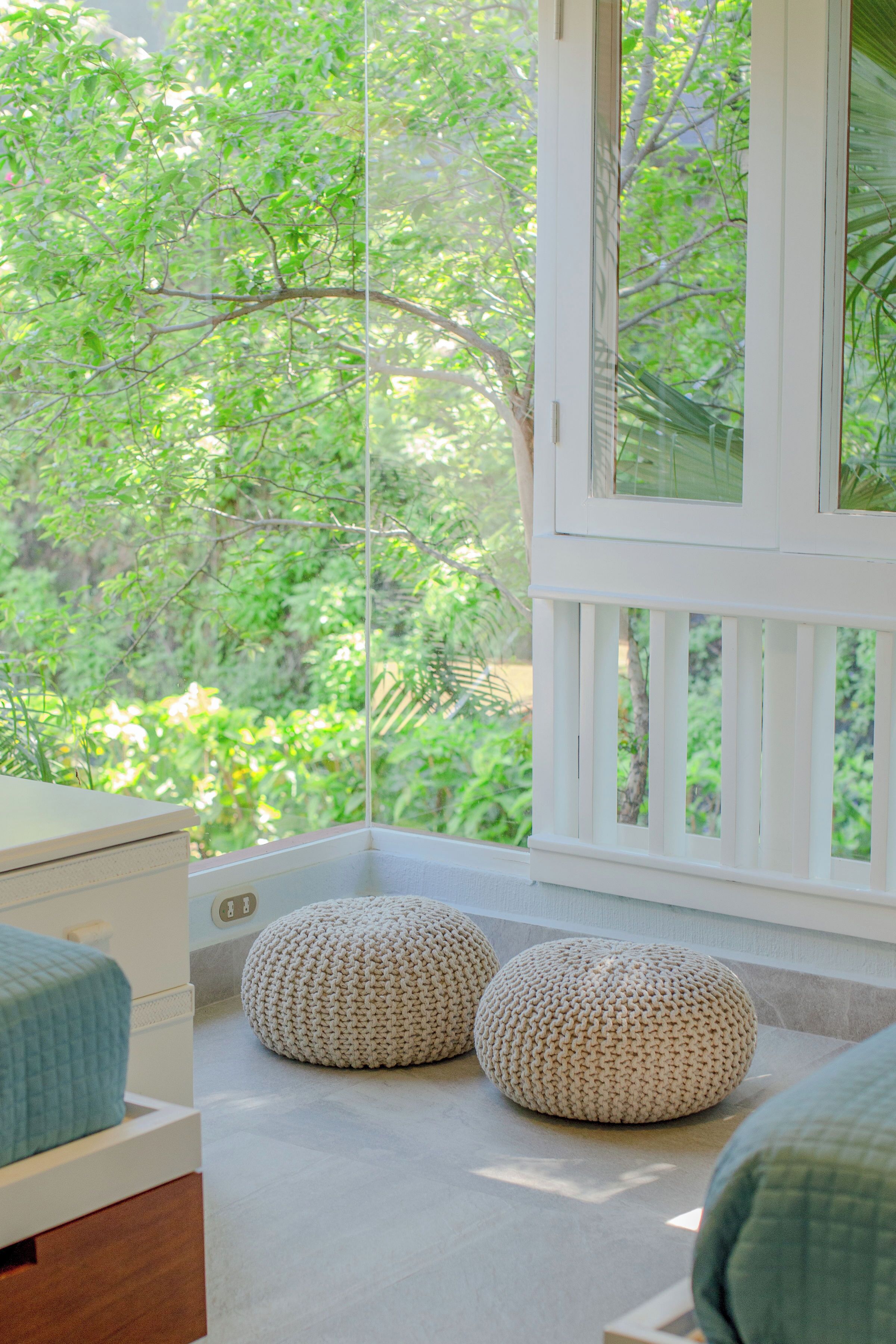 A room with a large window, a bed, two round poufs, and a wooden nightstand.