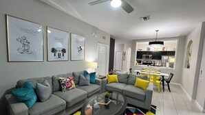 A modern living room with a gray sofa, yellow cushions, and a colorful geometric rug. There are framed Mickey Mouse prints on the wall, a ceiling fan, and a kitchen in the background. - ToyStory - Poolside/Water park Gound Level next to Disney (Kissimmee)