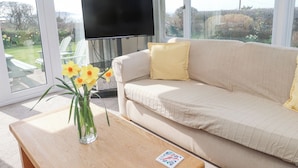 Un salón con un sofá color beige, un televisor de pantalla plana, una mesa de centro con un jarrón de flores y vistas a un jardín a través de grandes ventanales.