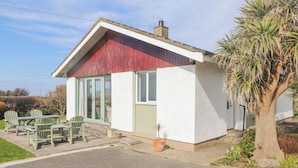 Una casa pequeña de un solo piso con techo rojo y paredes blancas, que cuenta con un patio con sillas verdes y una mesa.