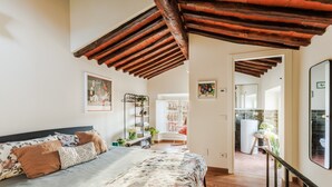 Une chambre avec un plafond en bois, un lit recouvert d’une literie à motifs floraux, une chaise blanche et une petite table sur laquelle est posée une plante.