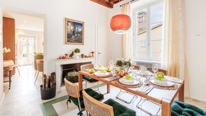 Un coin repas avec une table en bois dressée pour six personnes, entourée de chaises en osier avec des coussins verts. Une cheminée avec un manteau de couleur blanche est visible au fond, et une grande fenêtre offre une vue sur un bâtiment à l’extérieur.