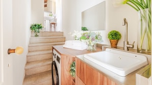 Une salle de bain moderne avec un meuble-lavabo en bois, un lavabo blanc et un miroir au-dessus. Un escalier mène à une autre pièce, et une plante en pot est posée sur le meuble-lavabo.