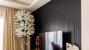 A modern living room with a flat-screen TV mounted on a black wall, a green console table with decorative items, and a large white flower arrangement in a black pot. - Apartment in El Sheikh Zayed (Giza Governorate)