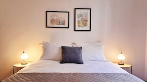 Un lit bien fait, recouvert d’une courtepointe matelassée, avec deux oreillers et un coussin décoratif. Deux lampes de chevet sont placées de chaque côté du lit. Deux œuvres d’art encadrées sont accrochées au mur au-dessus du lit.