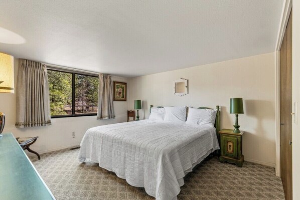 Un dormitorio con una cama grande, dos mesitas de noche con lámparas, una ventana con cortinas y vistas a árboles.