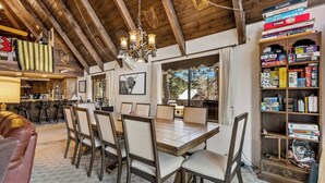 A dining area with a wooden table and chairs, a chandelier, and a bookshelf filled with books and games.