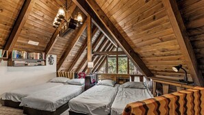 A cozy attic bedroom with two beds, wooden beams, a bookshelf, and a window providing a view of the outdoors.