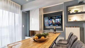 A modern living room with a wooden dining table, a comfortable grey sofa, a flat-screen TV mounted on the wall, and a small shelf with decorative items.